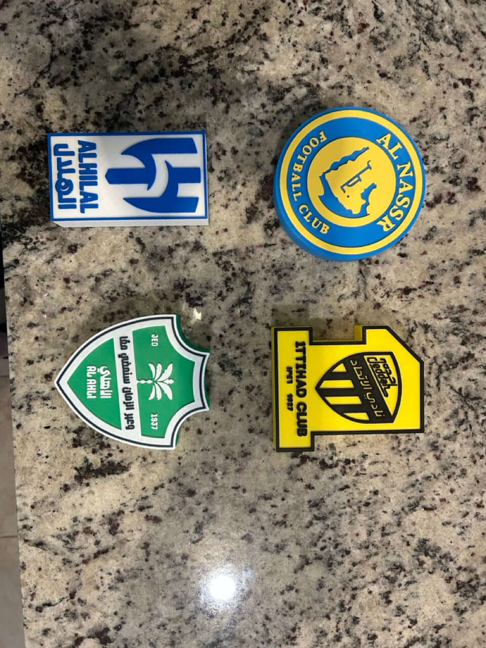 Collection of colorful football club badges on a granite surface, featuring Al Ahli, Al Nassr, and others.