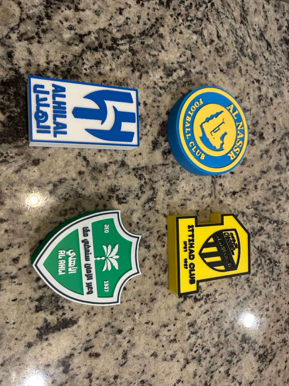 Four colorful 3D printed football club emblems on a granite surface, representing Al-Ahli and Al-Nassr teams.