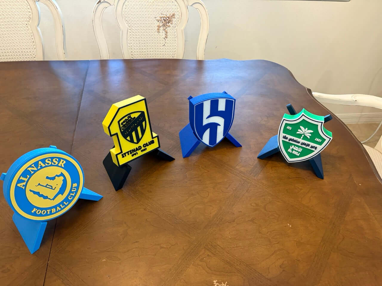 4 colorful 3D printed plaques of Al-Nassr, Al-Ittihad, Al-Hilal, and Al-Ahli football clubs on a table.