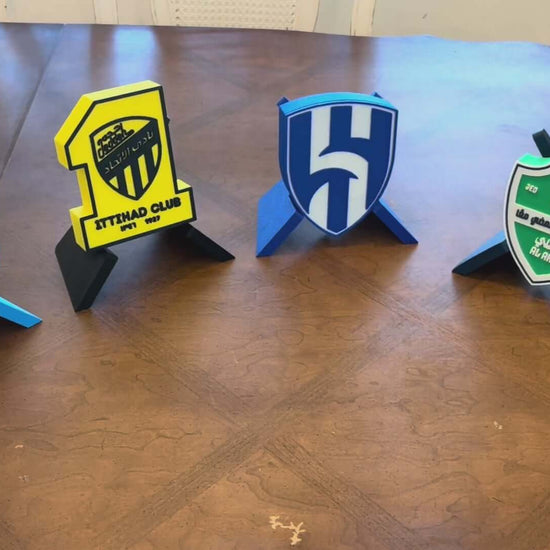 3D printed plaques of Saudi football clubs: Al-Nassr, Ittihad, Al-Hilal, and Al-Ahli on a wooden table.