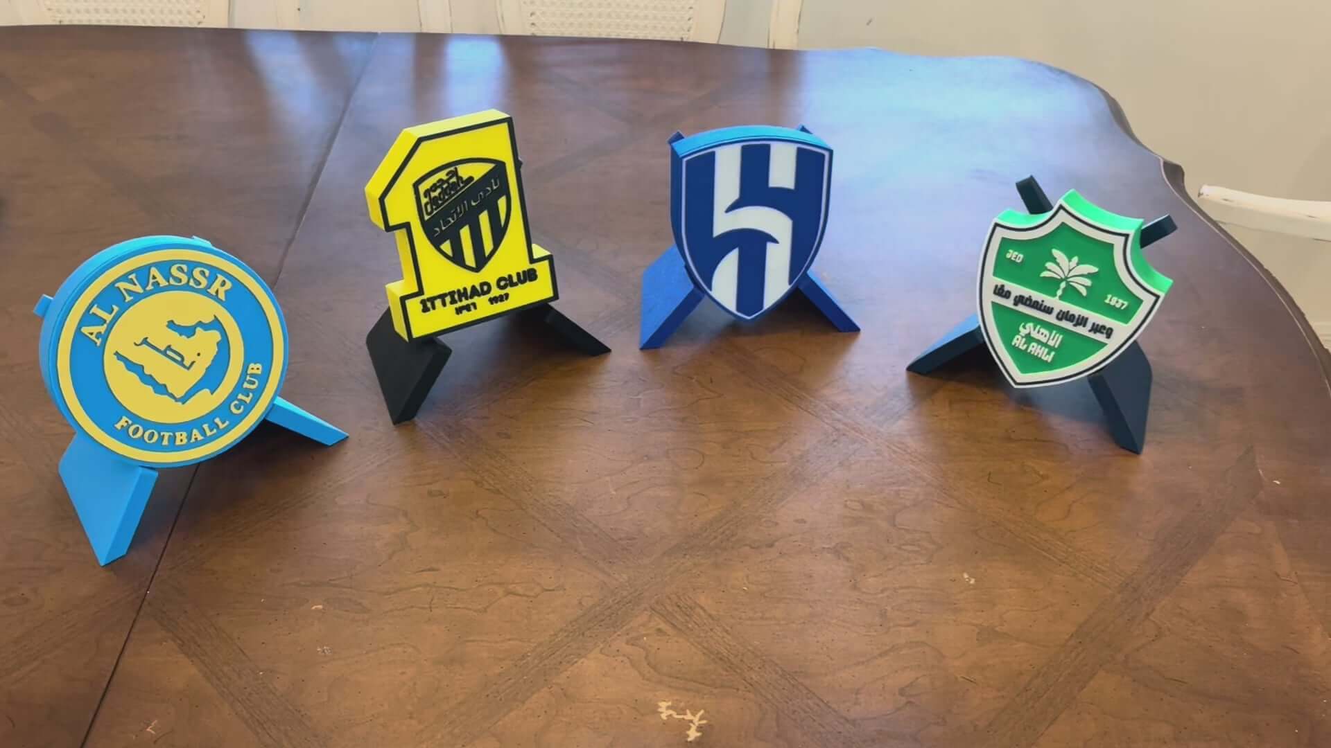 3D printed plaques of Saudi football clubs: Al-Nassr, Ittihad, Al-Hilal, and Al-Ahli on a wooden table.